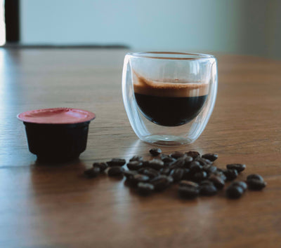 A table with roasted Arabica coffee beans a double wall espresso cup filled with an espresso shot showing thick crema.  A Central Bru proprietary espresso coffee capsule with red lid 