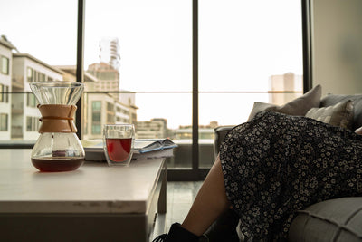 Woman sitting on couch with coffee table in front of her, on the coffee table is a Central Bru hourglass dripper for pour over coffee and a double wall glass filled with coffee.  