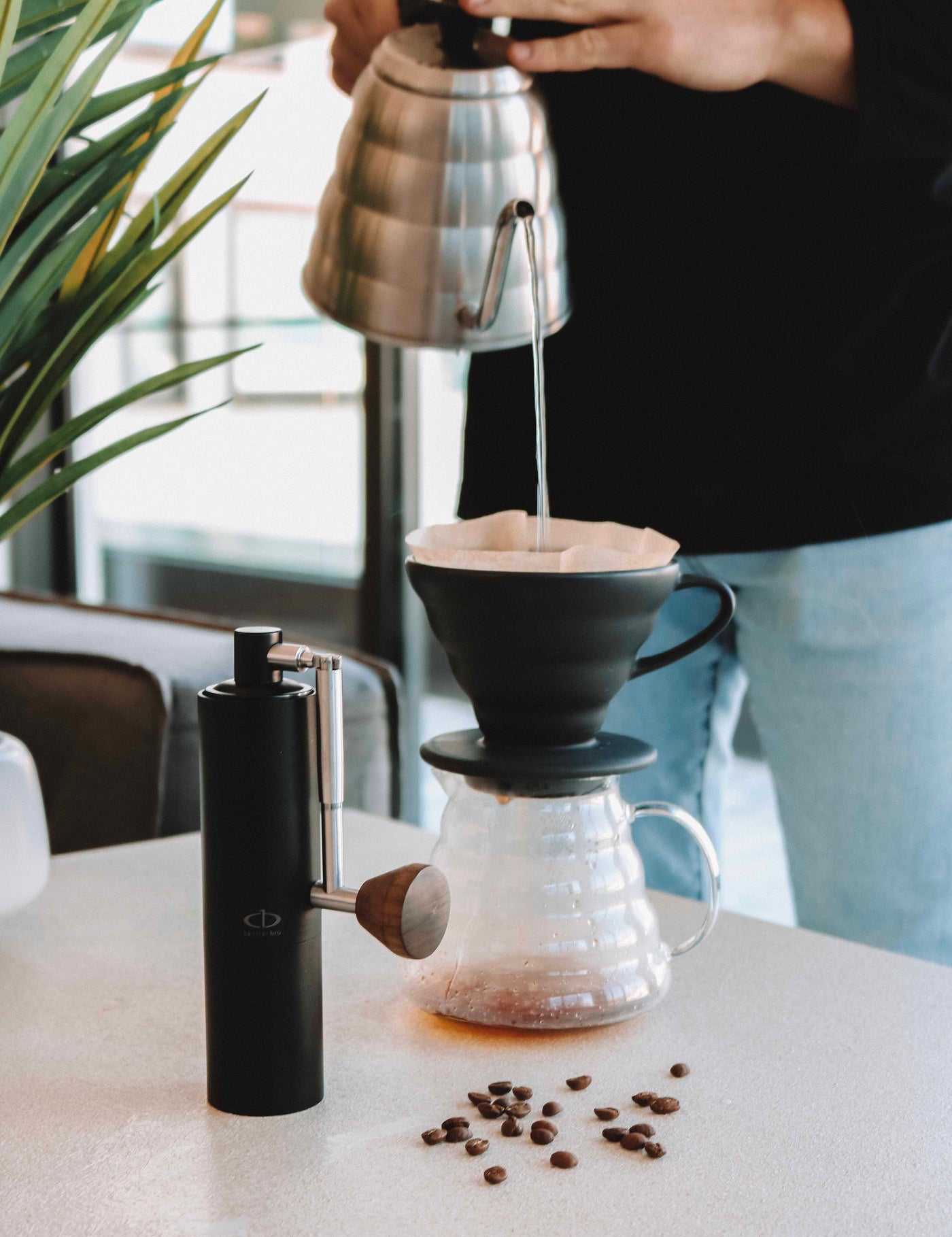 A Central Bru black anodized aluminum coffee grinder with silver hand and wood knob.  Man pouring water with a stainless steel gooseneck kettle in a v60 making a pour over coffee that is filtering into a glass coffee server