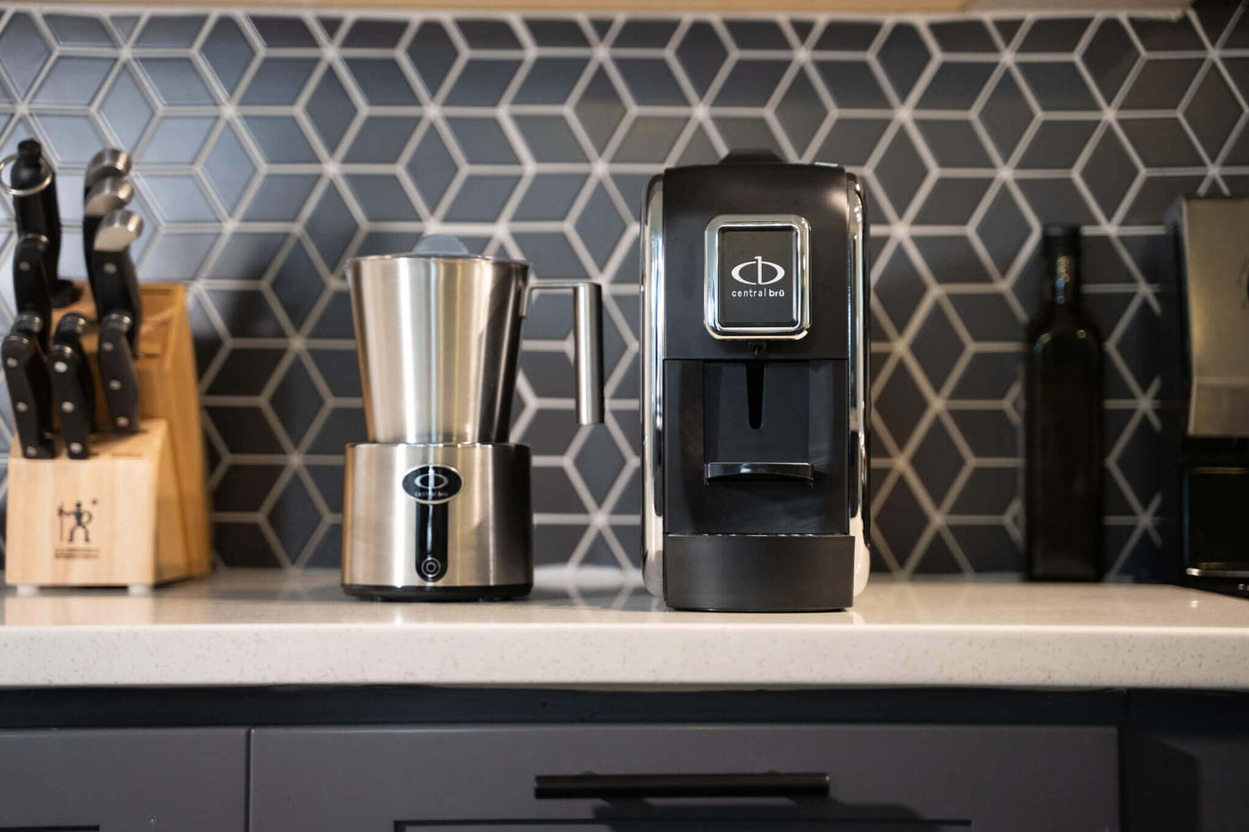 Central Bru espresso brewing system with Central Bru milk frother on a white kitchen counter with grey cabinetry all compliment each other as the espresso brewing system is matte black with chrome sides and the milk frother is full stainless steel