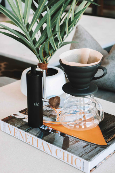 A Central Bru coffee grinder, black anodized aluminum with silver handle and wood knob on a coffee table with a v60 pour over and coffee server all sitting on top of a book