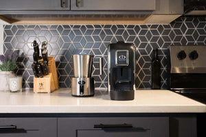 Central Bru espresso brewing system with Central Bru milk frother on a white kitchen counter with grey cabinetry all compliment each other as the espresso brewing system is matte black with chrome sides and the milk frother is full stainless steel